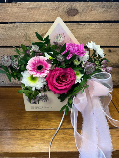 Love Letter - A charming floral keepsake presented in an elegant envelope-style container, filled with vibrant pink tones, crisp whites and lush greenery. Designed to capture the feeling of a heartfelt message, this arrangement combines soft textures, delicate details and a graceful ribbon finish for a romantic and contemporary look. Perfect for birthdays, anniversaries, thank-you gifts or simply letting someone know they are in your thoughts, this delightful design delivers beauty and sentiment in equal measure.