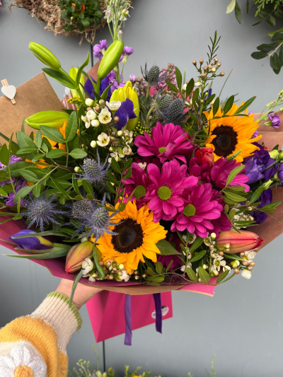 Fiesta - Bursting with bold colour and natural charm, this eye-catching hand-tied bouquet brings together rich jewel tones, sunny accents and fresh seasonal textures. Designed to capture the carefree beauty of a wildflower meadow, it features a lively mix of striking blooms, delicate details and lush greenery for a joyful, uplifting display. Wrapped in coordinating papers and finished with elegant ribbon, this bouquet is perfect for birthdays, celebrations, congratulations or simply brightening someone's day with a cheerful splash of colour.