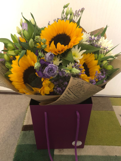 Sunflowers - Radiating warmth and happiness, this vibrant hand-tied bouquet combines rich golden tones with soft lilac accents and fresh seasonal greenery. Full of texture, colour and natural charm, it creates a cheerful display that instantly brightens any room. Beautifully presented in stylish wrapping and a coordinating gift bag, this uplifting arrangement is perfect for birthdays, celebrations, congratulations or sending a thoughtful burst of sunshine to someone special.